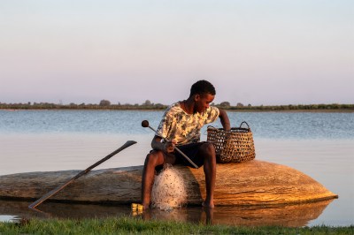 A young boy sitting on a log by a river, examining a basket next to him, with a paddle resting beside him.